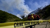 Close up shot of yellow tape stretched with the words: "CRIME SCENE DO NOT CROSS." Behind this barrier are three people dressed in yellow jackets and helmets with red backpacks. They are looking out at smoke billowing from the place where two green mountains meet against a blue sky.